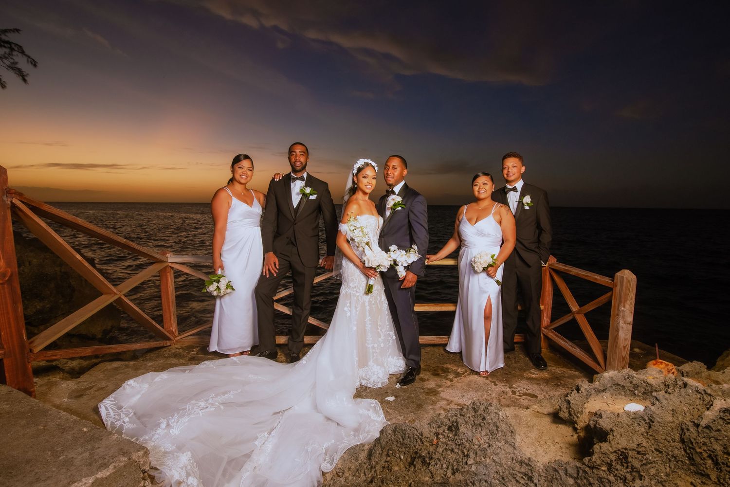 Wedding party poses on rustic wooden deck overlooking ocean at dusk.
