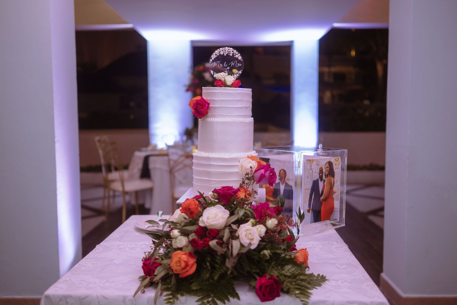 Elegant white wedding cake decorated with fresh flowers and purple uplighting.