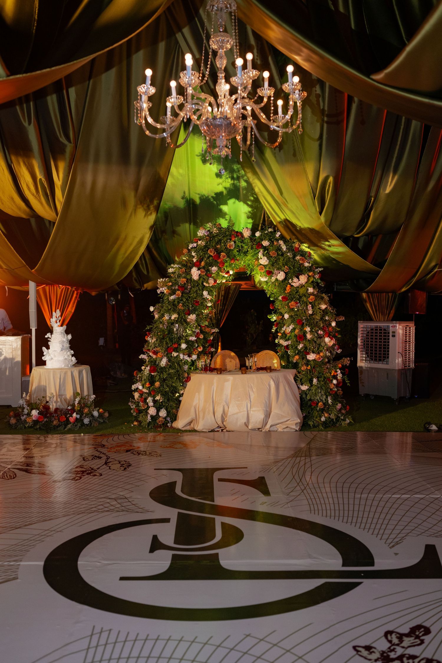 Elegant wedding reception venue with chandeliers, draped fabric, and floral arrangements on a patterned dance floor.