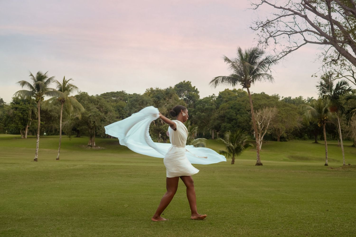 Dancing figure in white dress with flowing blue fabric in a lush tropical garden setting.