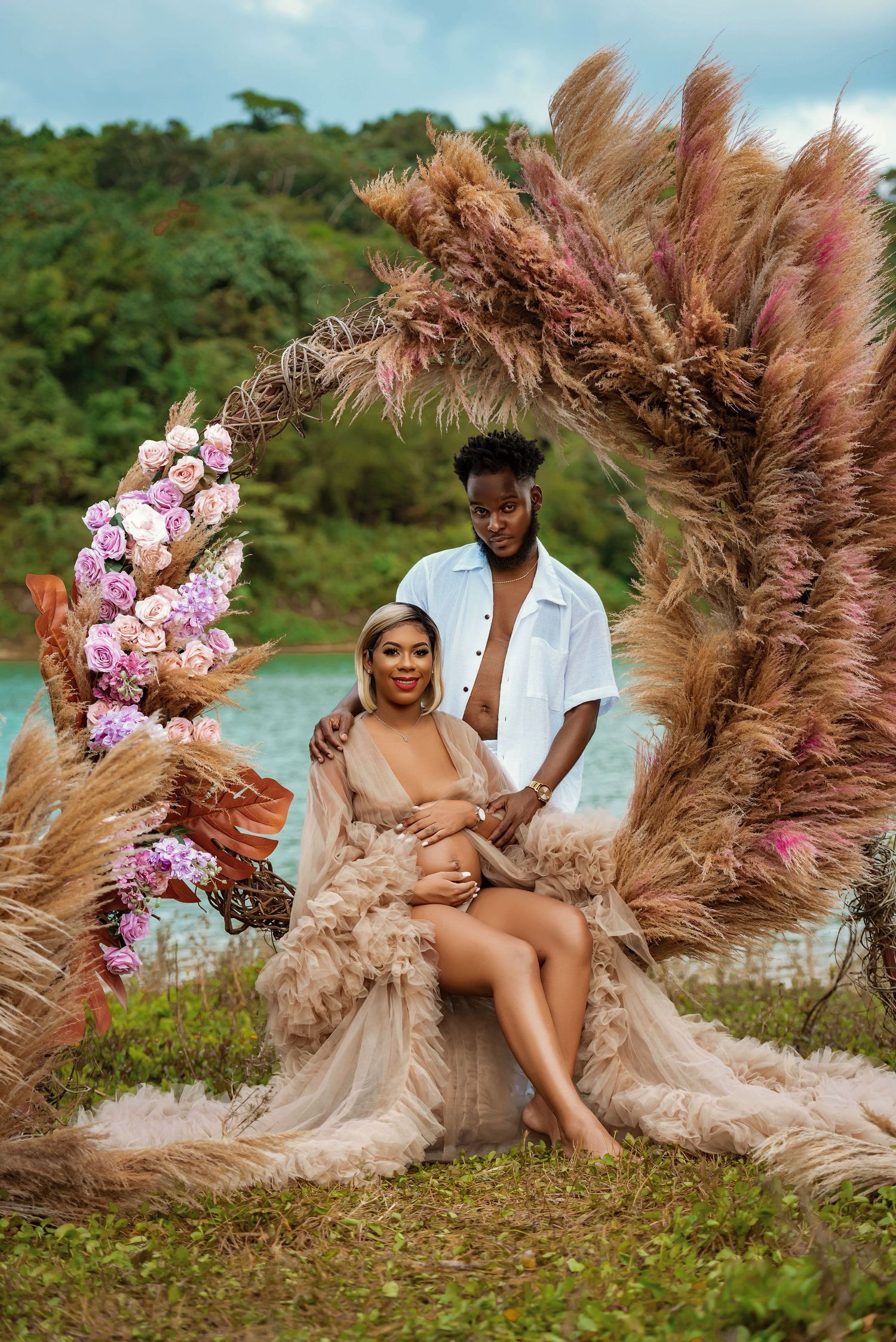Romantic couple poses under an elaborate pampas grass and orchid arch by the ocean.