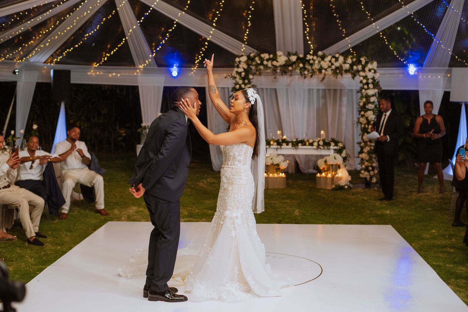 Wedding celebration on white dance floor under tent with string lights and floral decorations.