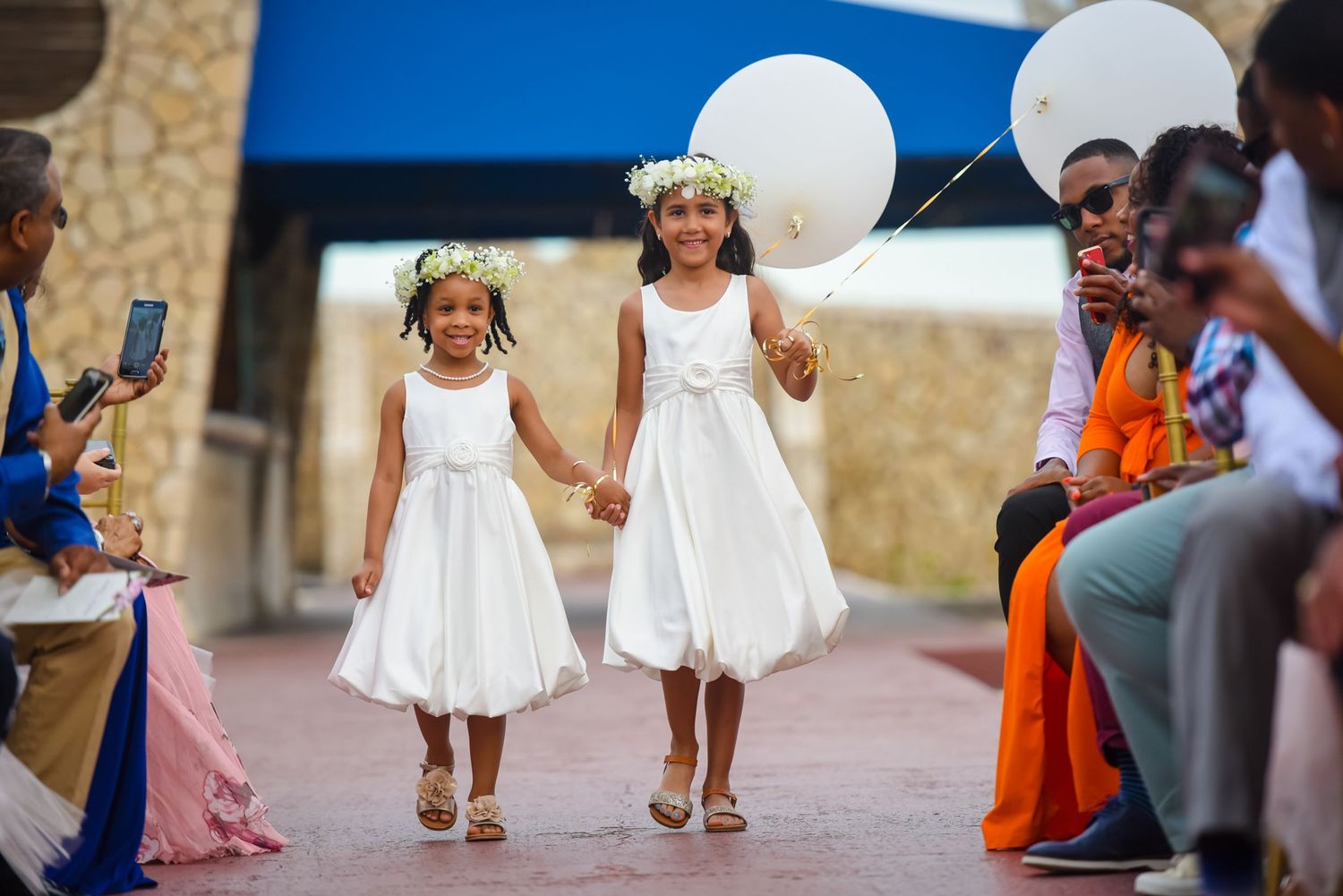 Two flower girls in white dresses carrying balloons down wedding aisle.