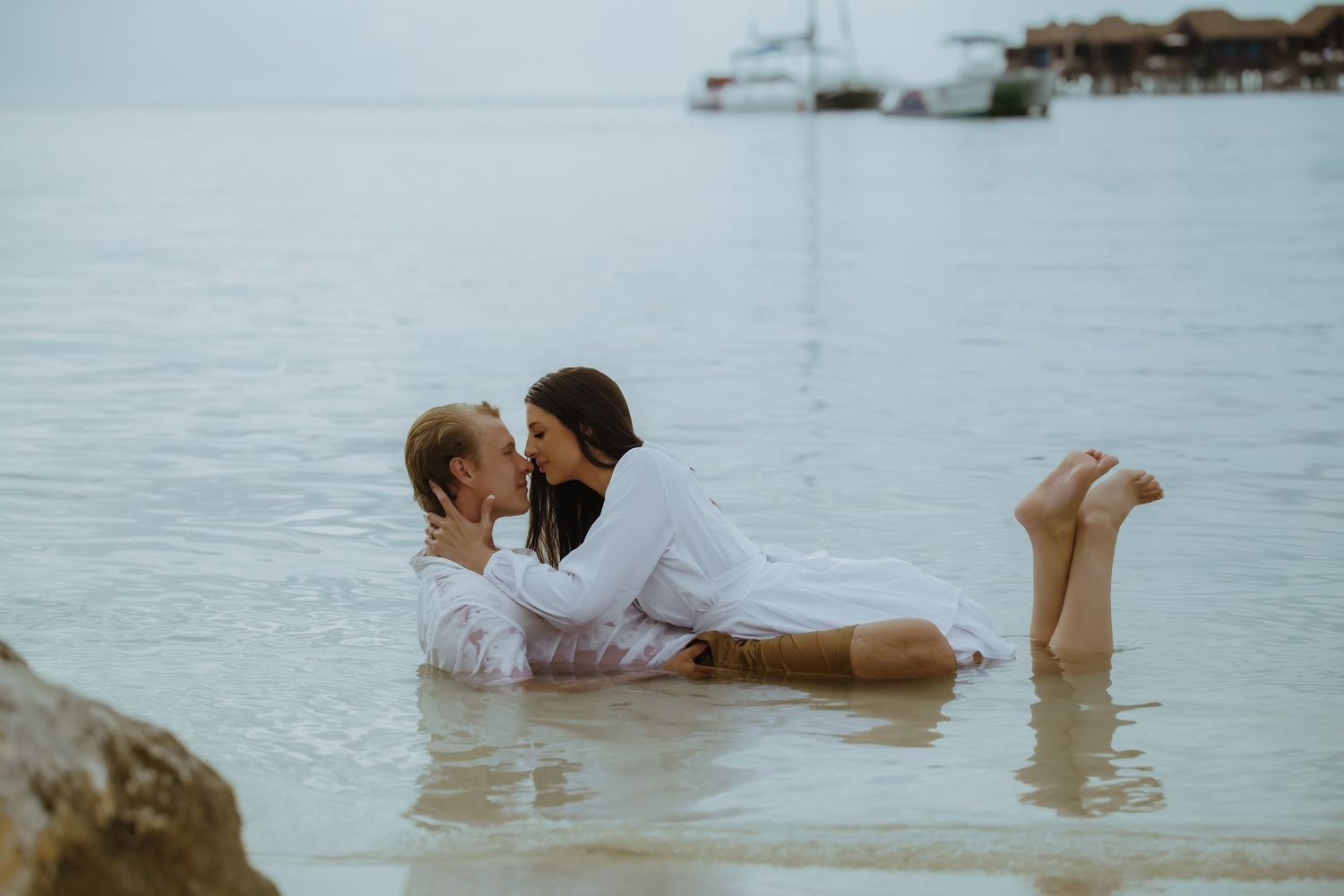 A couple in white clothing shares a romantic moment while lying in shallow ocean waters.