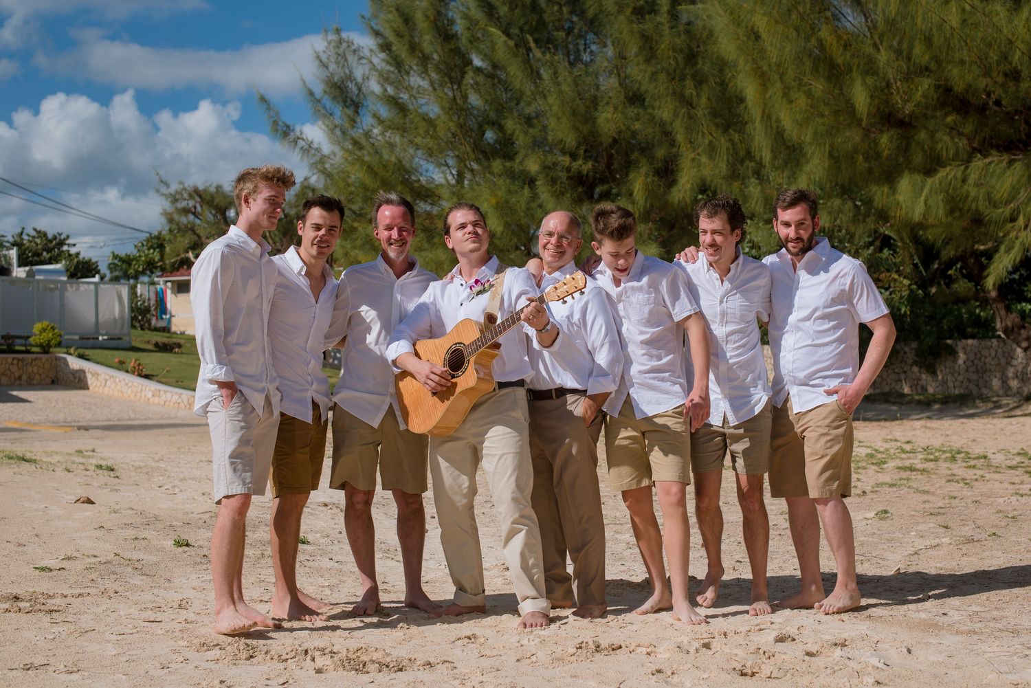 Wedding party members pose playfully on sandy beach wearing coordinated casual attire.