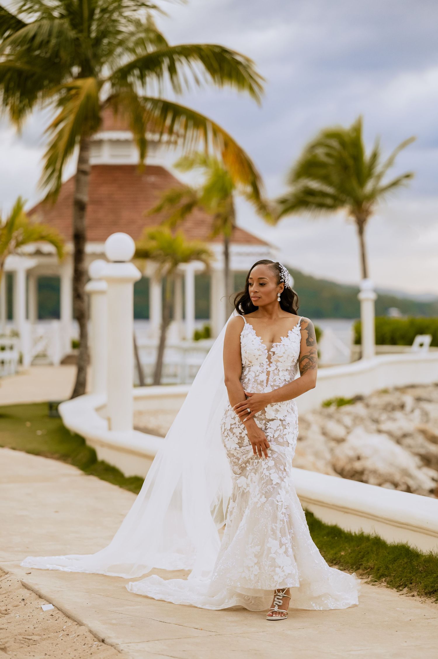 A bride in a lace wedding dress poses gracefully on a tropical waterfront path.