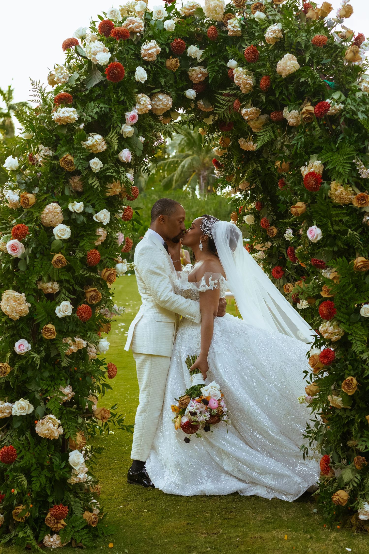 Wedding couple posing under a stunning floral arch with red and white roses in a garden setting.