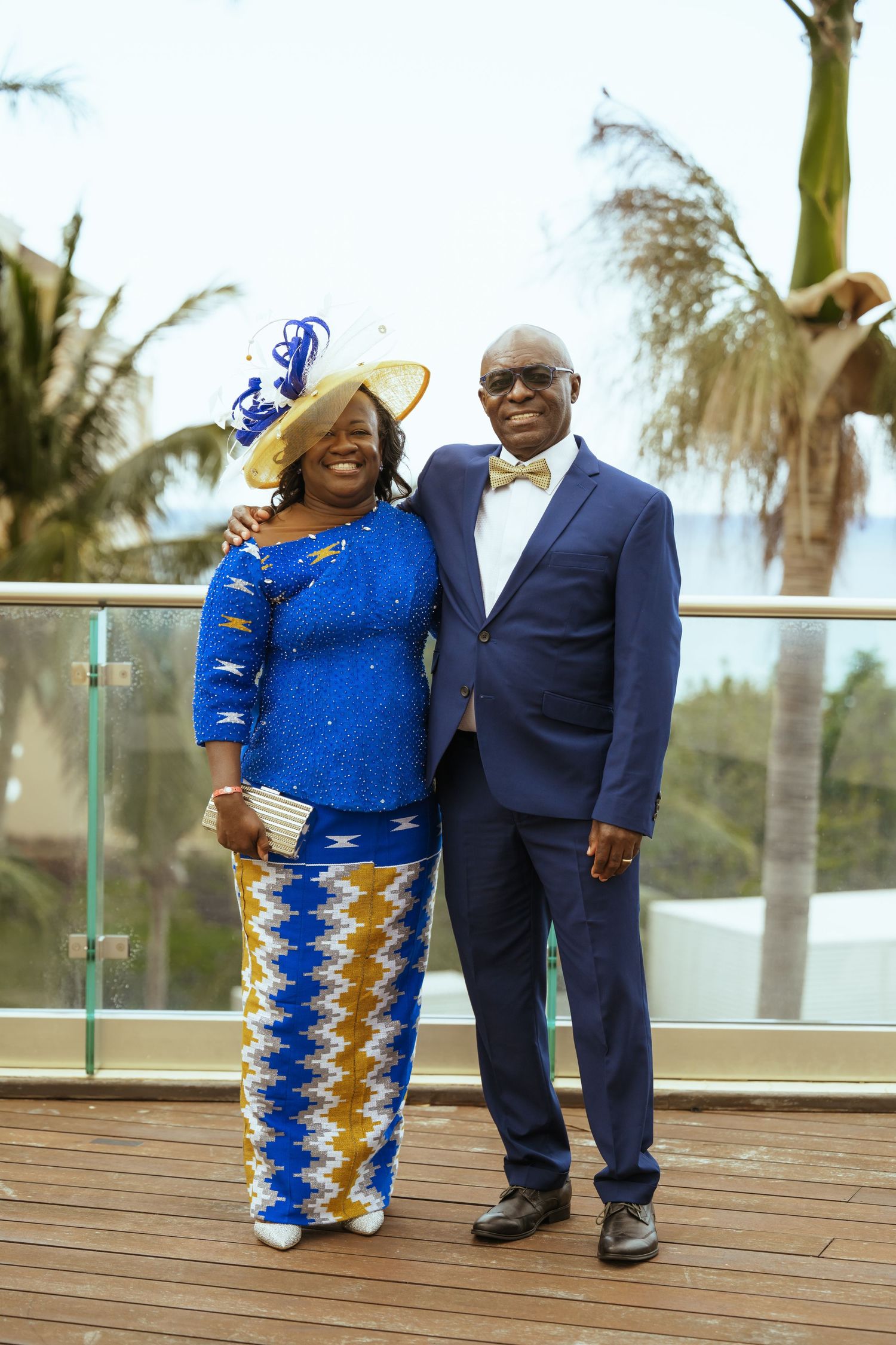 Couple in blue and navy formal attire pose together on wooden deck with palm trees in background.