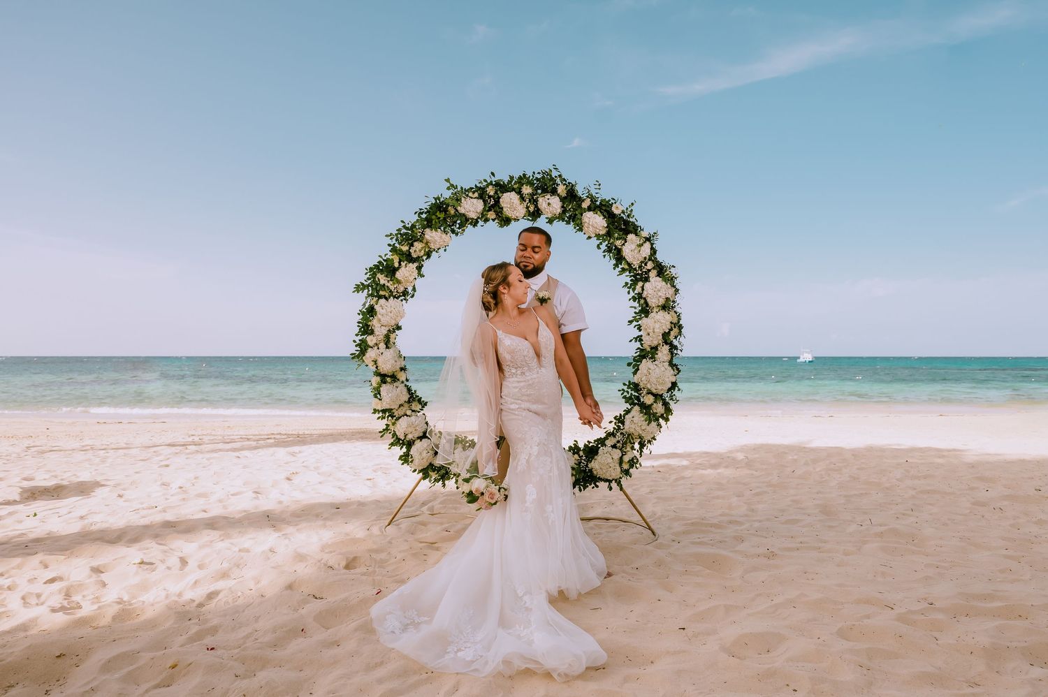 A romantic beach wedding ceremony setup with floral arch and chairs on white sand overlooking turquoise ocean waters.