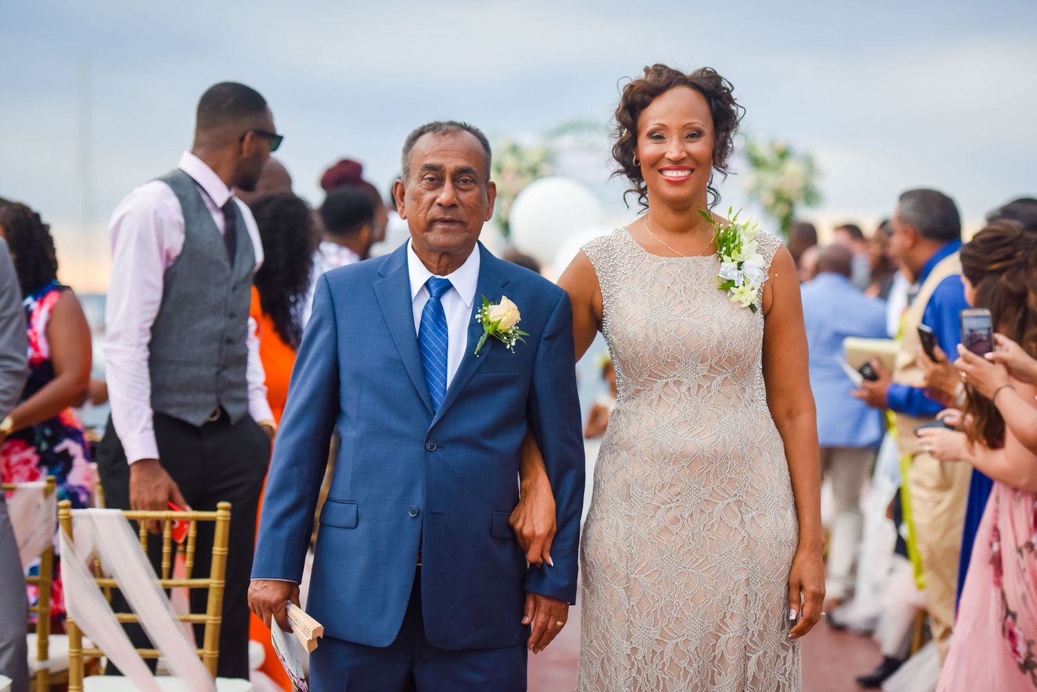 A couple walks arm in arm down a red carpet aisle during an outdoor wedding ceremony.