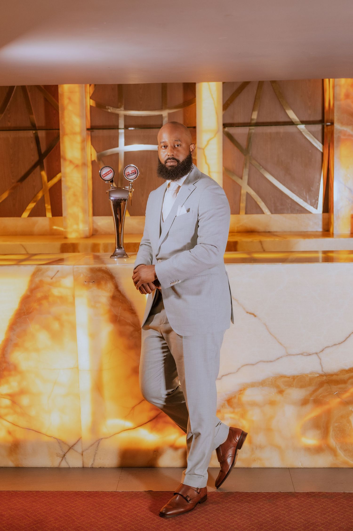 Stylish groom in light gray suit poses against warm-toned wall with artistic lighting effects.