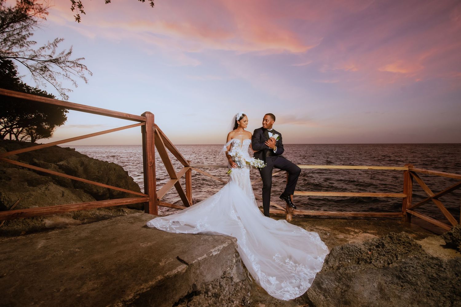 Dramatic sunset wedding portrait on cliff edge with ocean backdrop and wooden fence.