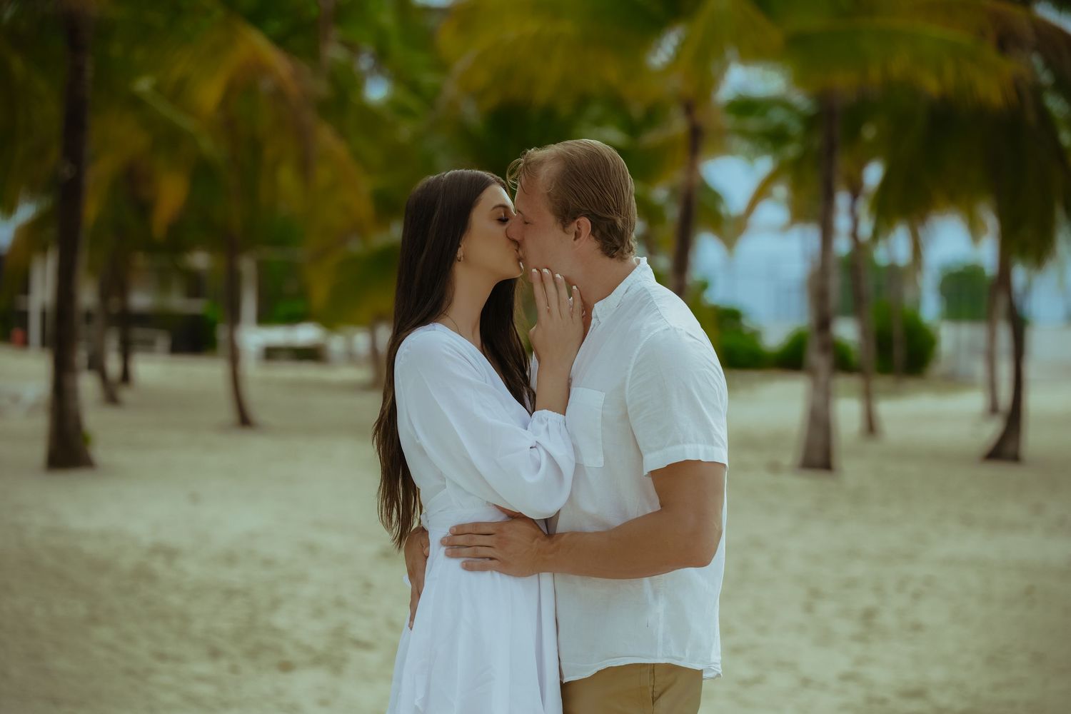 Romantic couple in white outfits share intimate moments on a tropical beach with palm trees in the background.