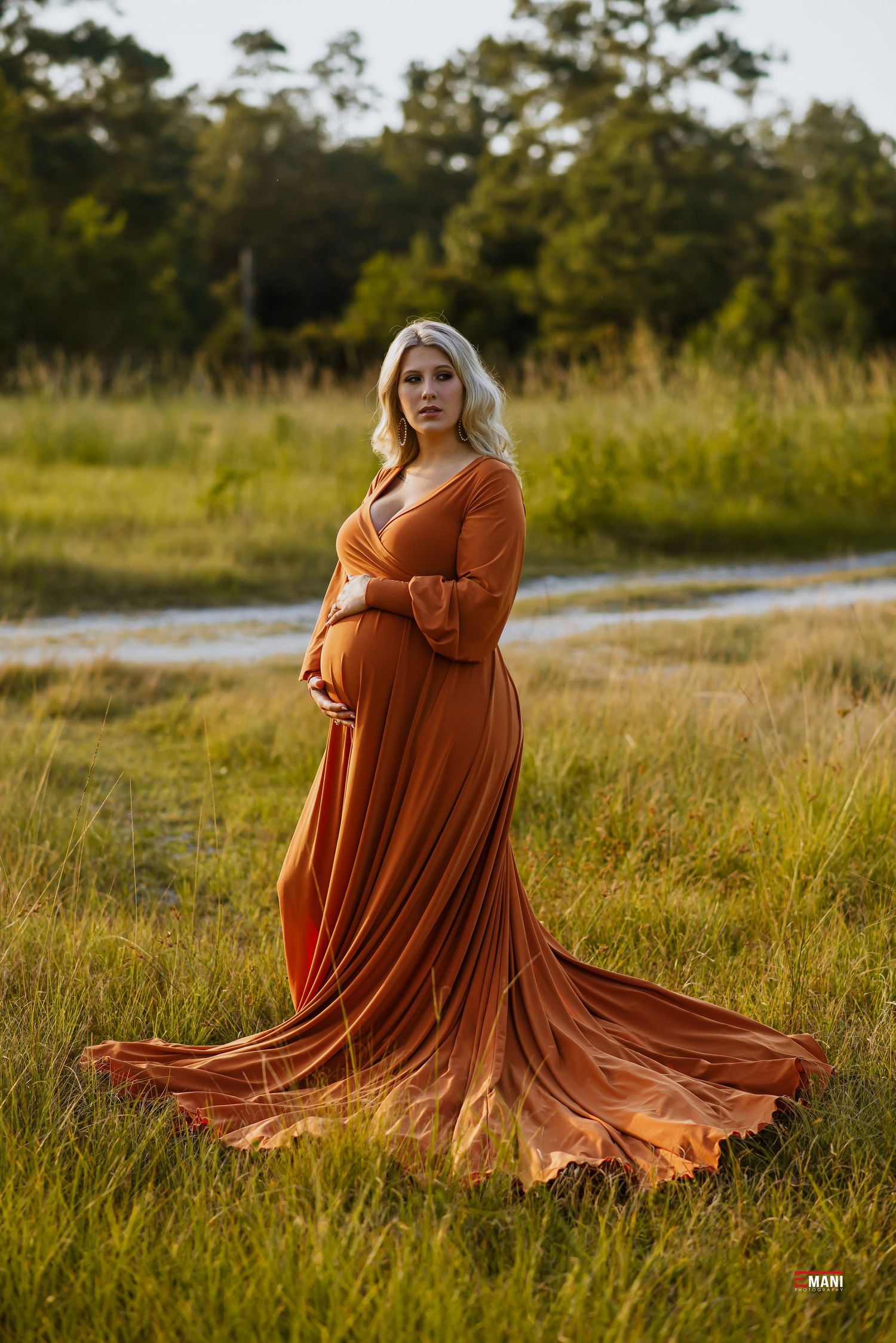 Expectant mother in flowing rust-colored dress poses in golden sunset field.