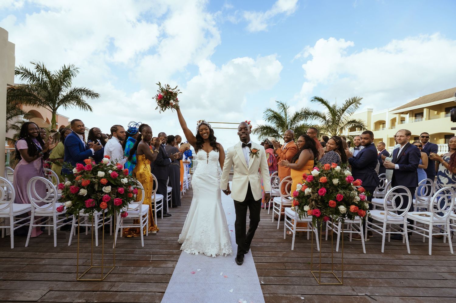 Beachfront wedding ceremony with palm trees and seated guests under cloudy skies.