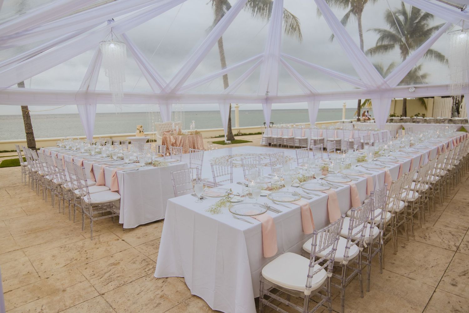Elegant beach wedding reception setup with long tables under a clear tent and palm trees in the background.