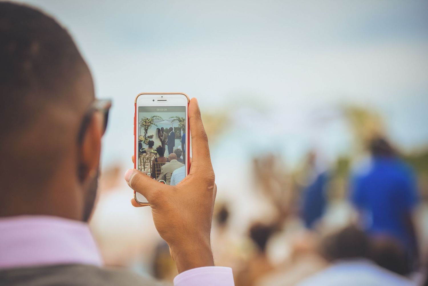 Guest captures wedding ceremony moments on smartphone during outdoor celebration.