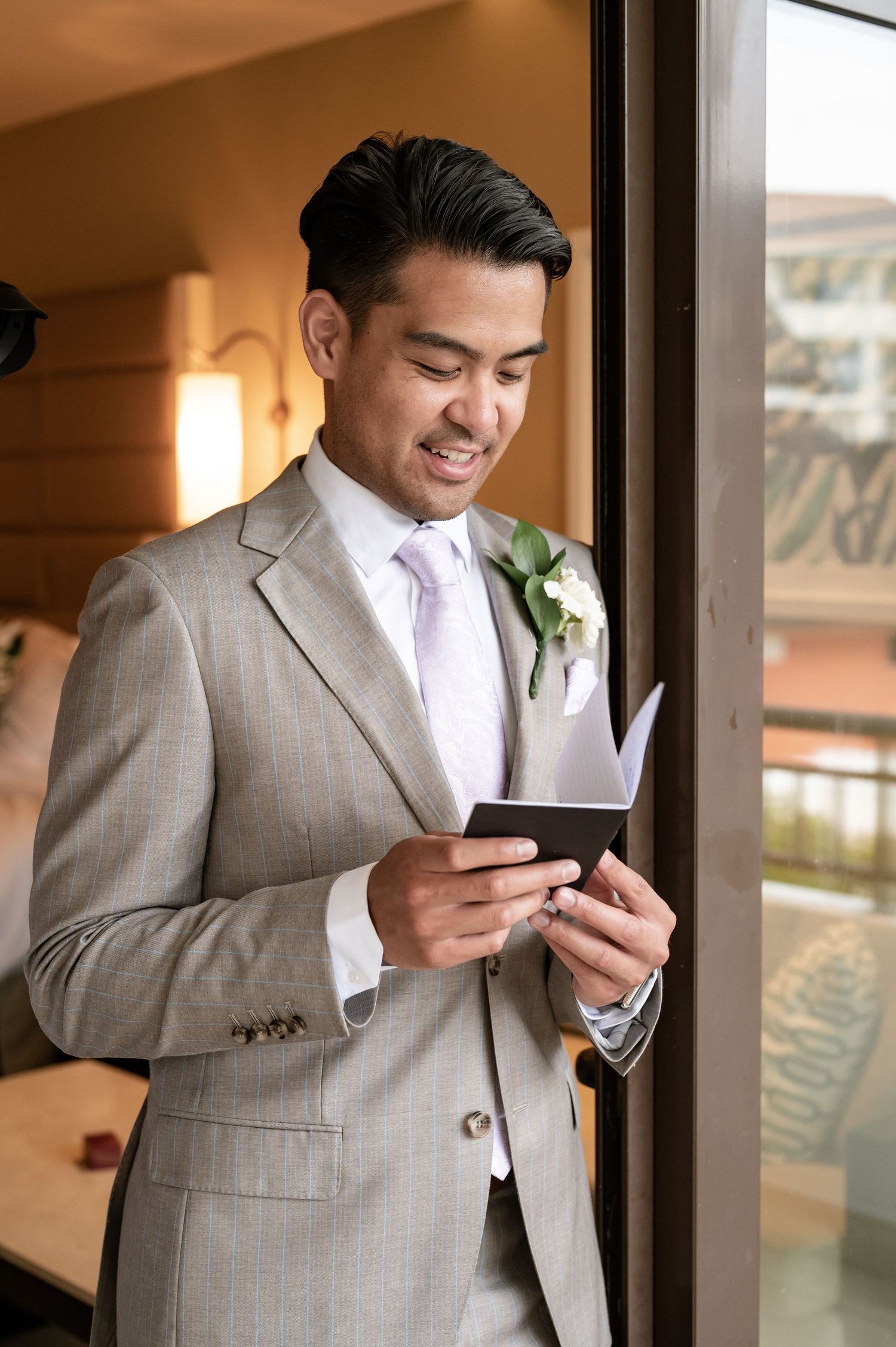 Groom in light grey suit reading personal vows by window.