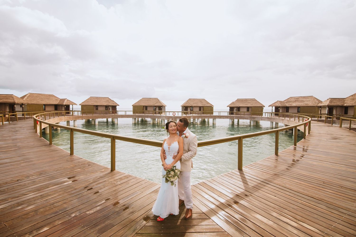 A couple poses romantically on a wooden boardwalk with overwater villas in the distance.