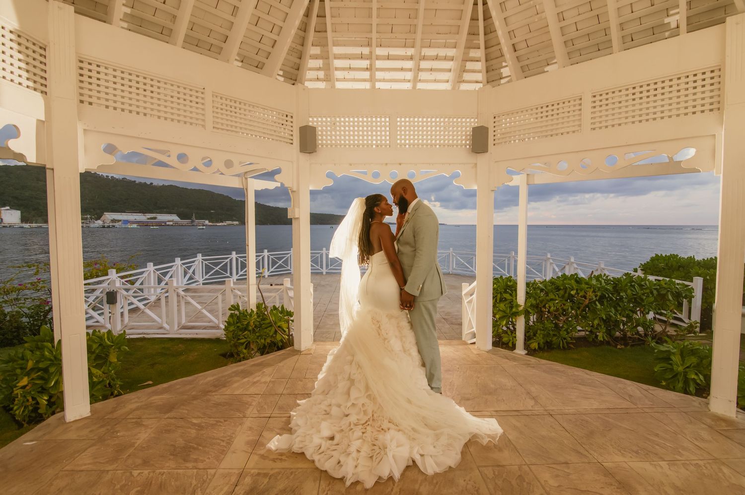 A wedding couple embraces under an elegant white oceanfront gazebo with panoramic sea views.