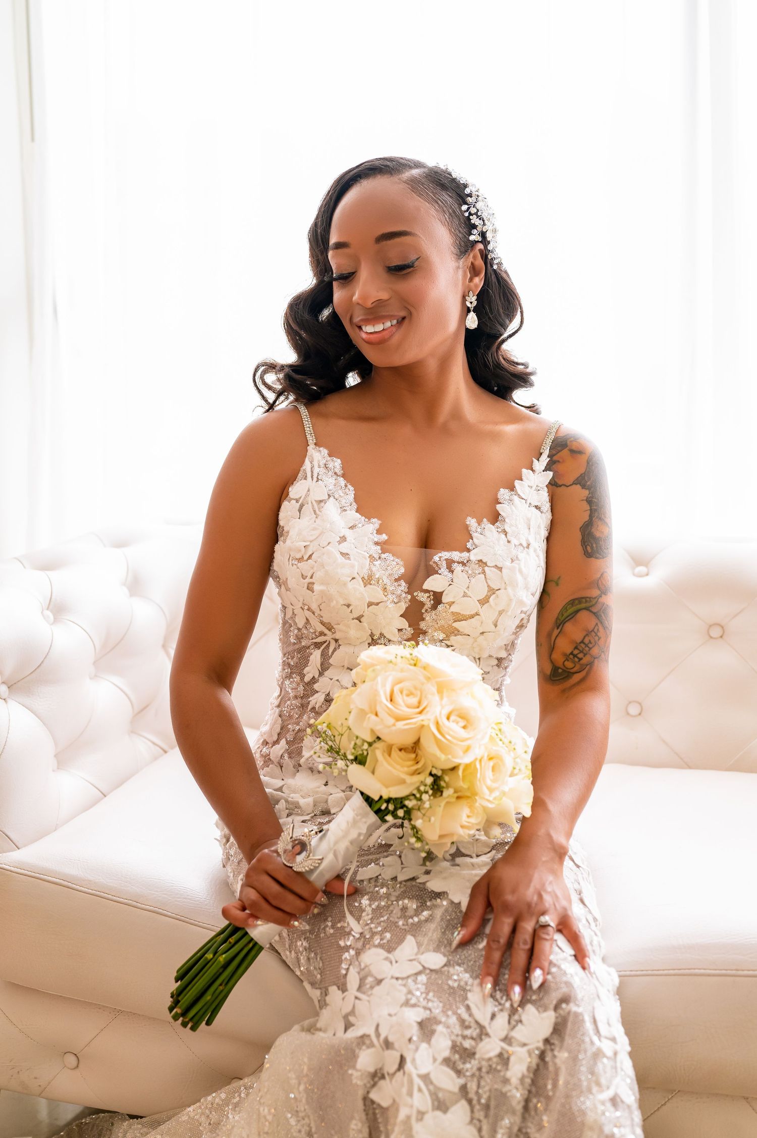 Bride in lace wedding gown holding cream roses while sitting on white couch.