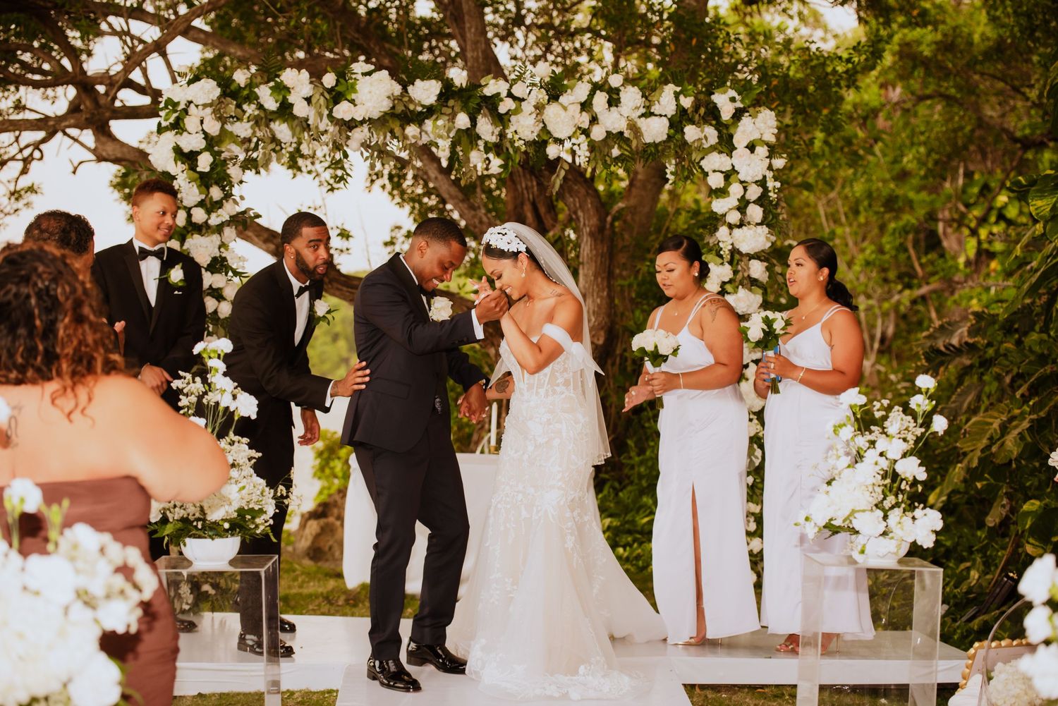 Outdoor wedding ceremony with white floral decorations and natural tree canopy.