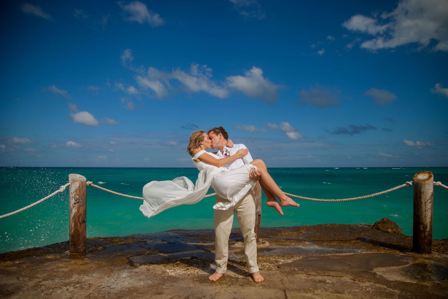 Romantic couple in white attire embracing on a tropical oceanfront walkway with turquoise waters in background.