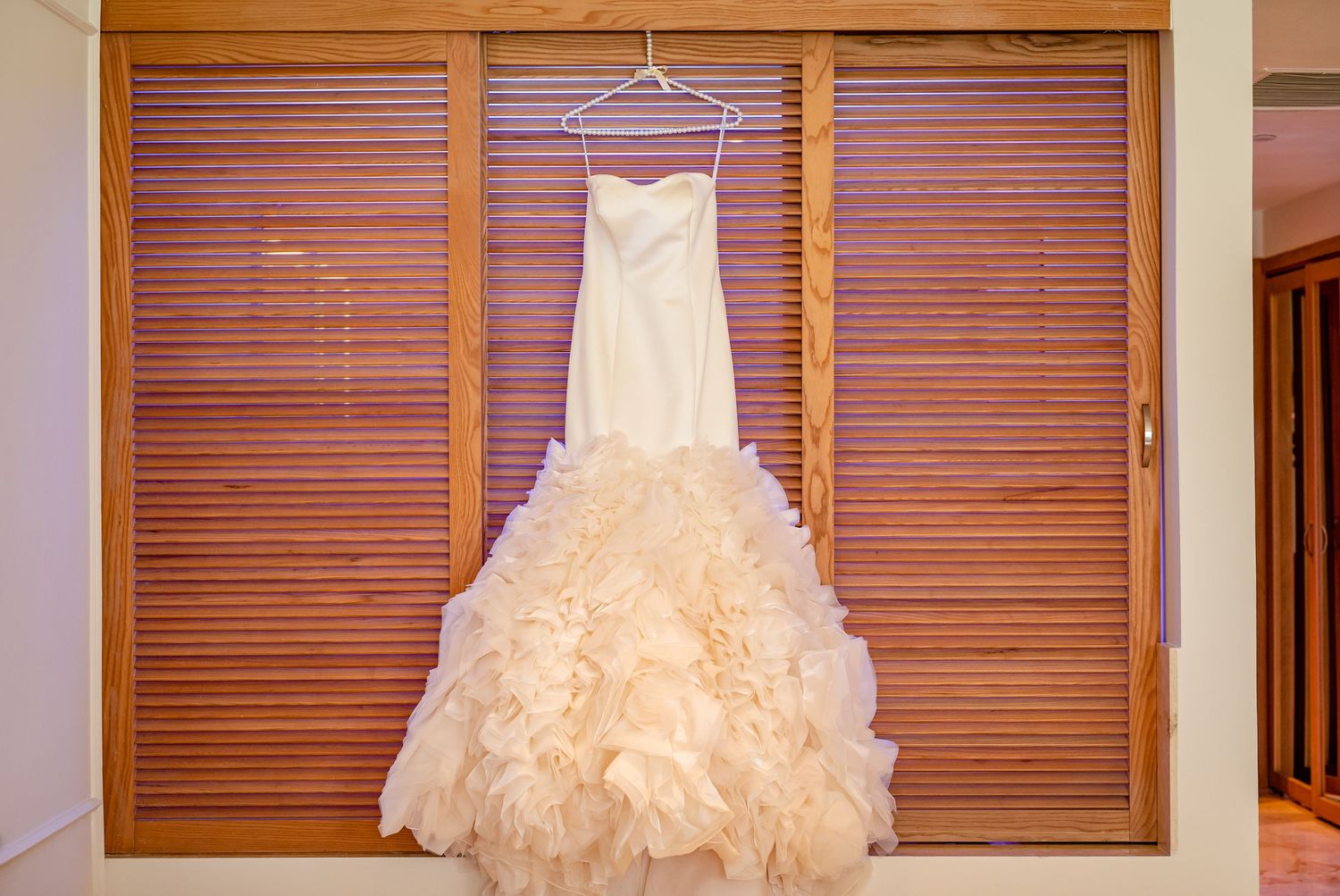 White ruffled wedding dress hanging against wooden shutters in natural light.