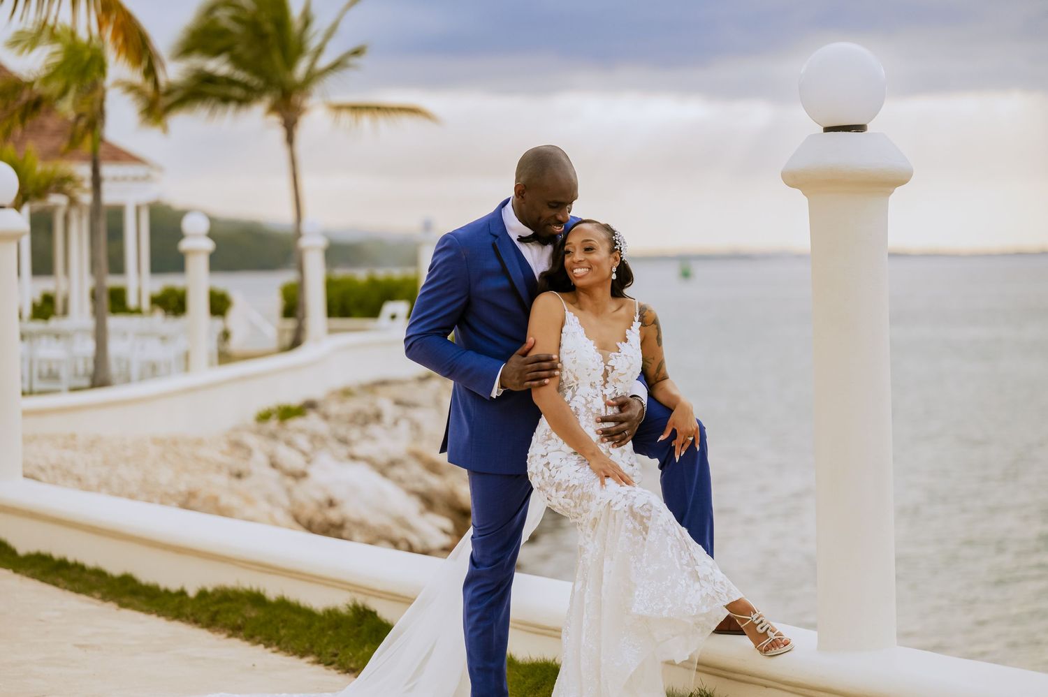 A couple in wedding attire embraces on a tropical waterfront promenade with palm trees.