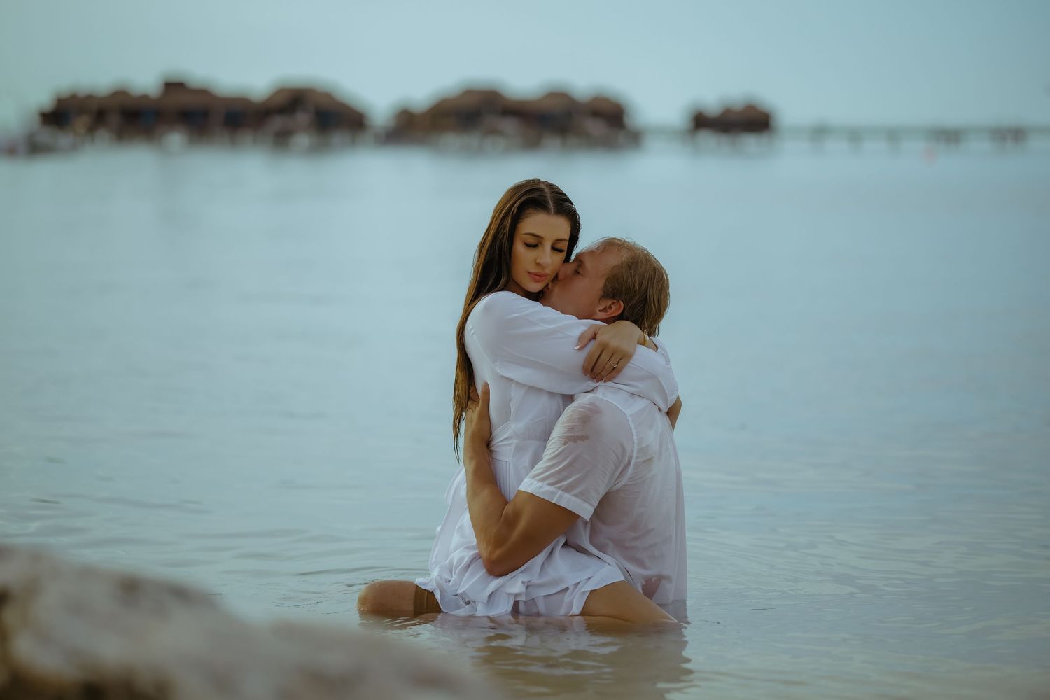 Romantic couple in white clothing embracing in shallow ocean water at sunset.