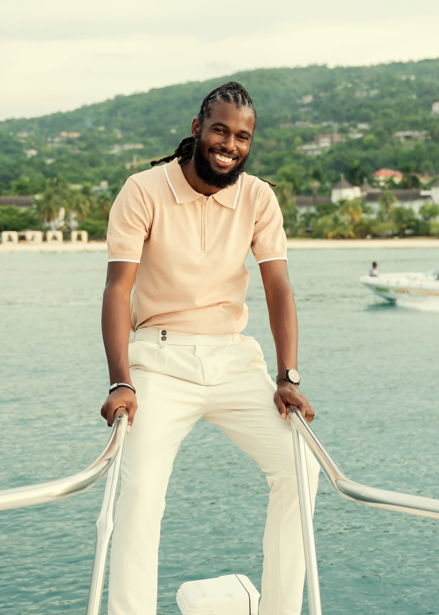 A person in a cream colored outfit poses against a scenic waterfront backdrop with boats and green hills.