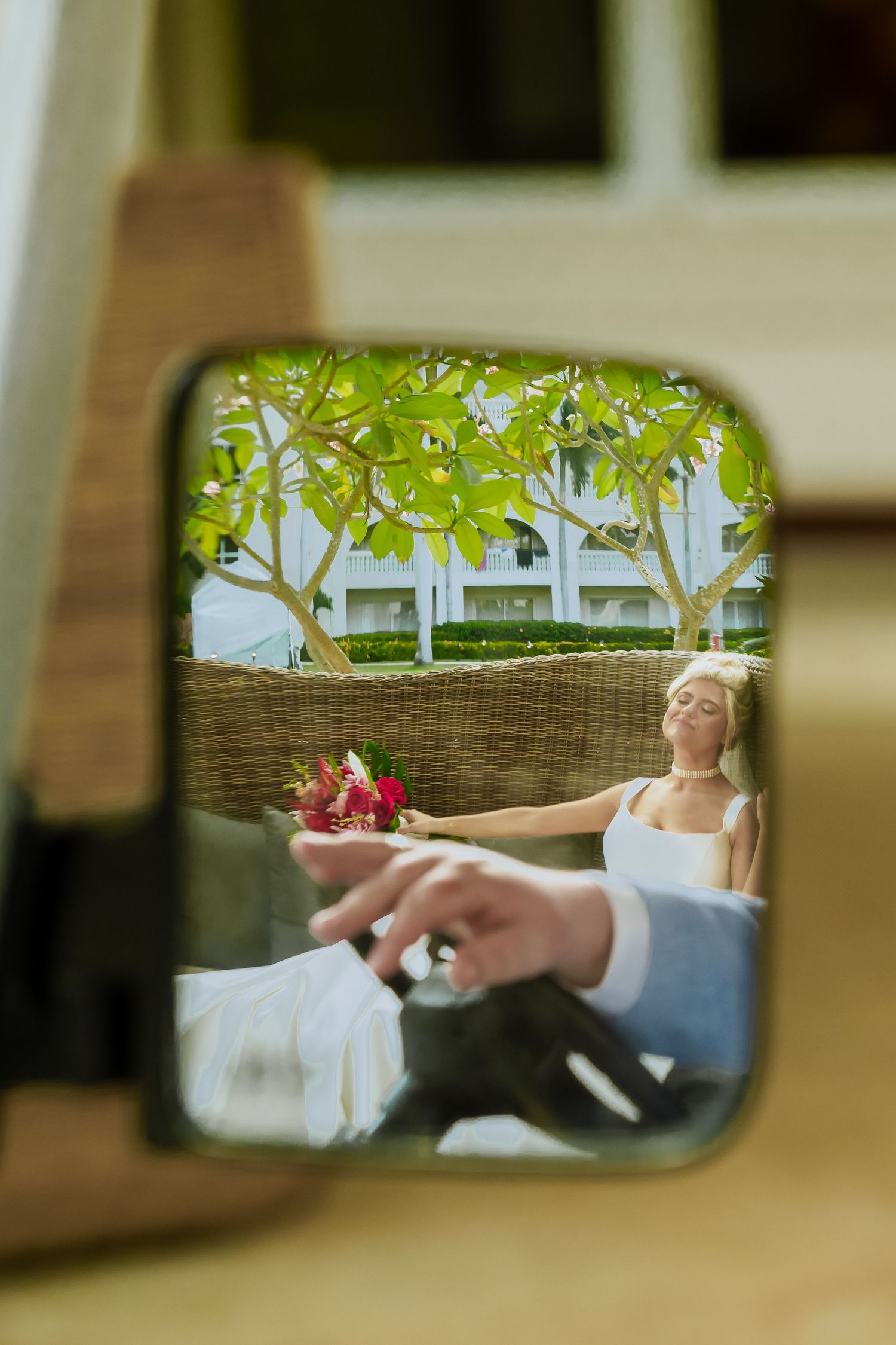 Creative car mirror reflection shot showing bride in white dress with tropical background.