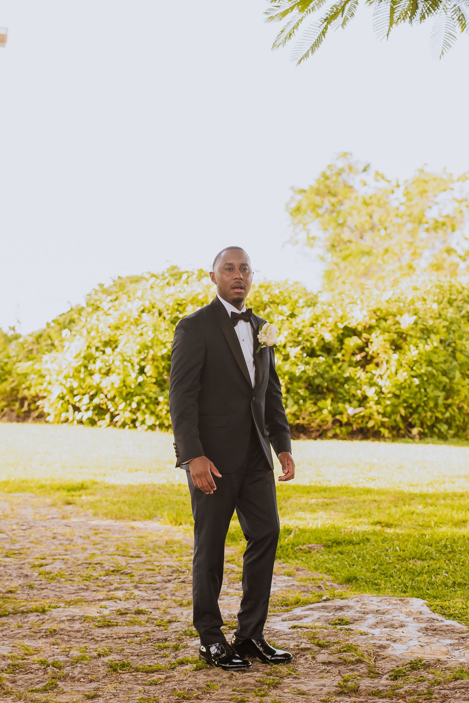 Person in formal black tuxedo posing outdoors in a garden setting.