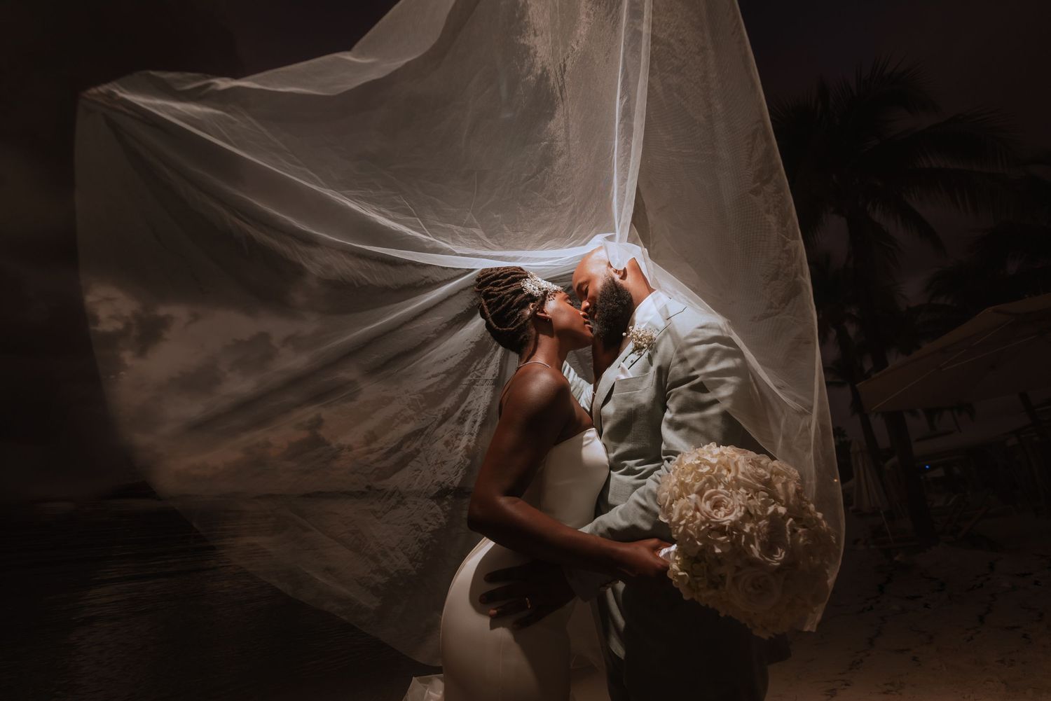 Dramatic silhouette of wedding couple with flowing veil against dark sky.