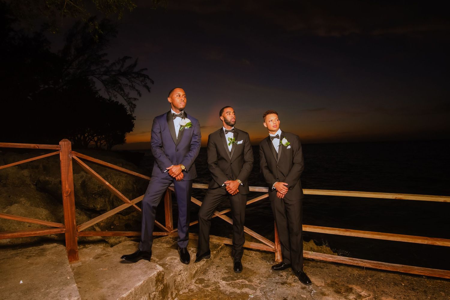 Three groomsmen in formal suits pose on wooden railing at twilight.