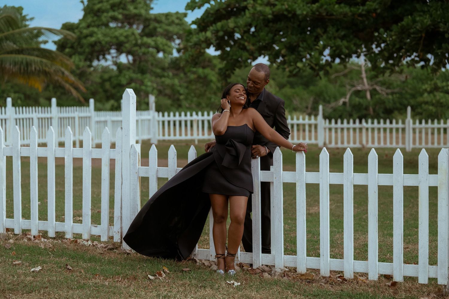 White picket fence stretches across a sunny lawn with trees in the background.