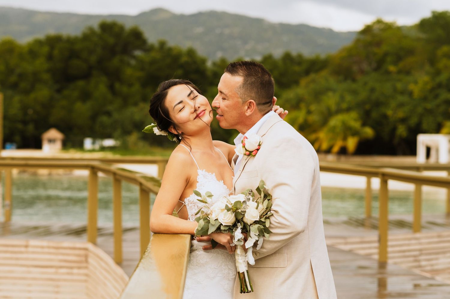 Couple embraces on a wooden dock with mountains and overwater bungalows in the background.