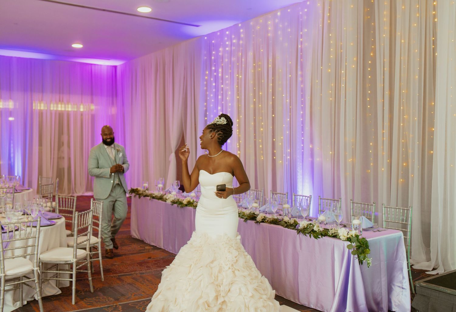 Wedding reception space decorated with white curtains, purple uplighting and floral arrangements.