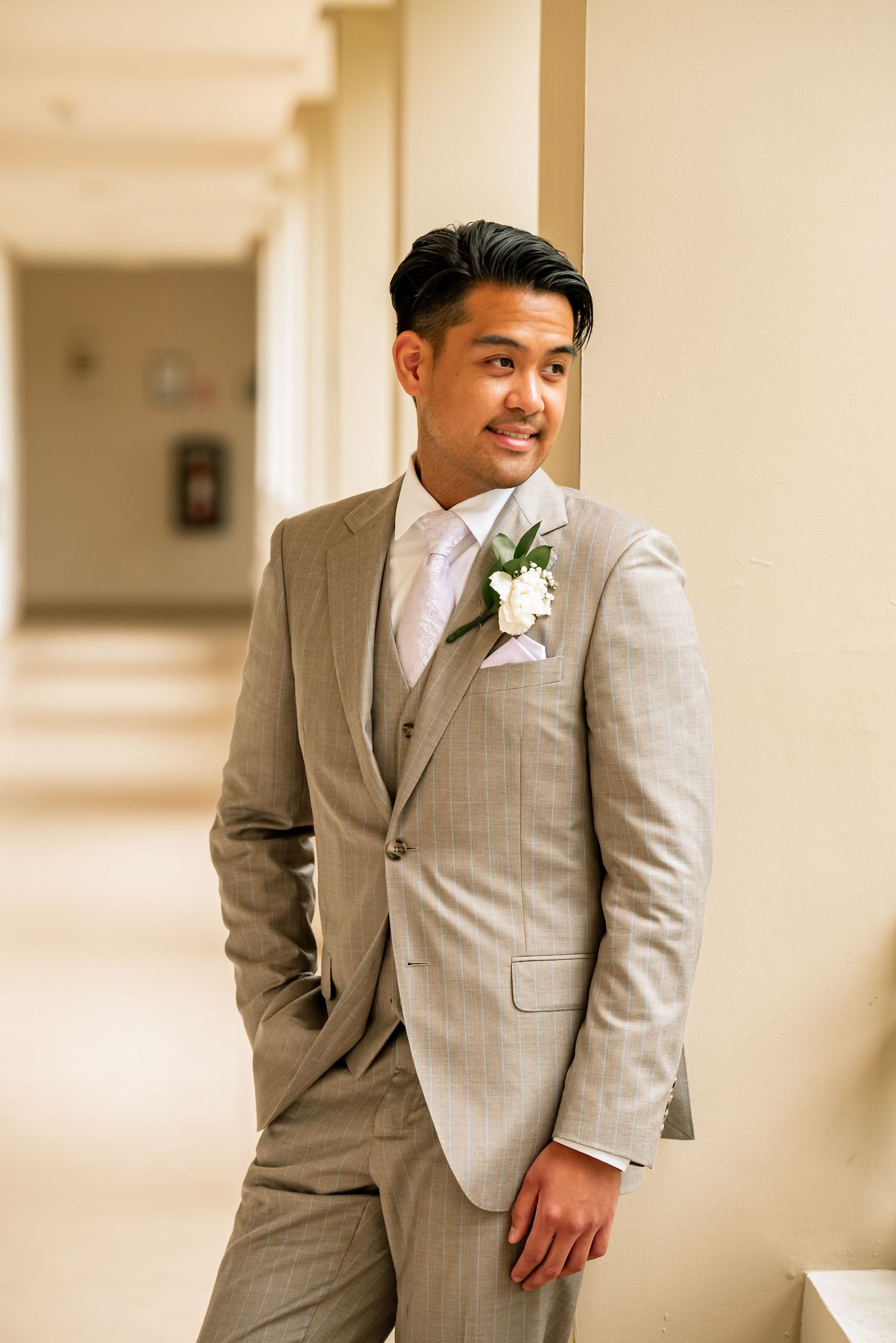 A groom in a light gray suit and white boutonniere poses in a bright hallway.