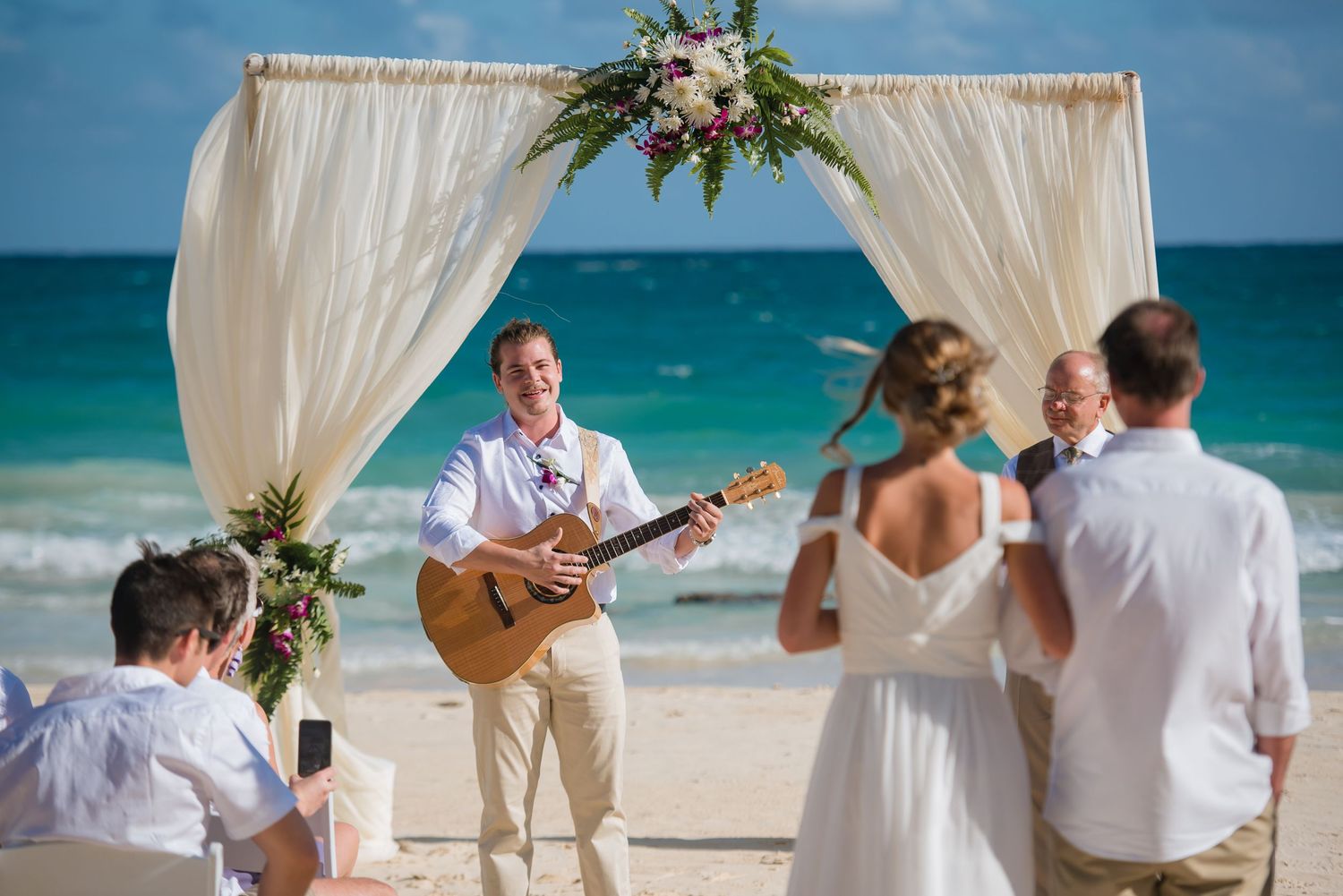 Intimate beach wedding ceremony under white fabric arch with tropical ocean views.