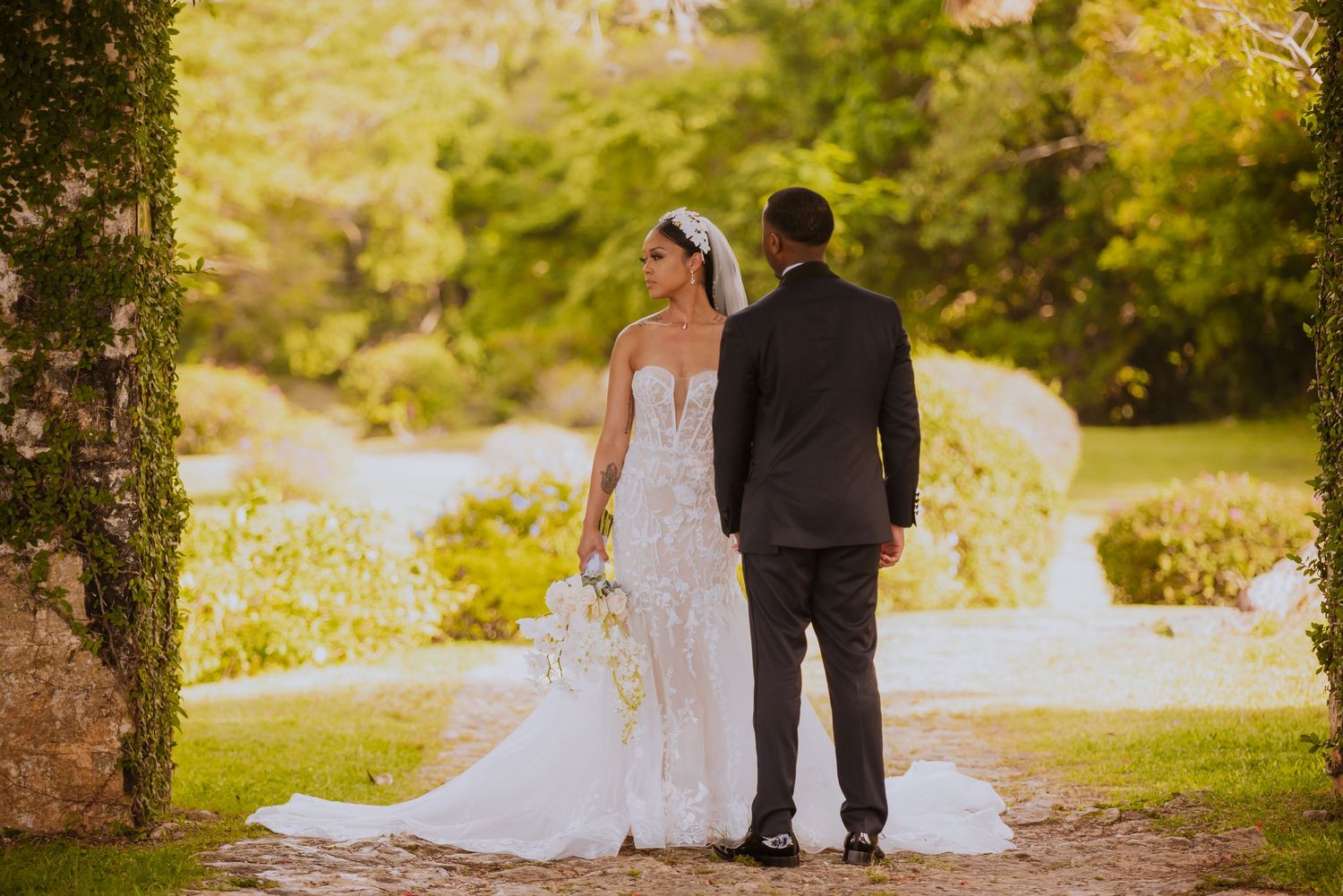 A wedding couple walks hand in hand along a tree-lined garden path.