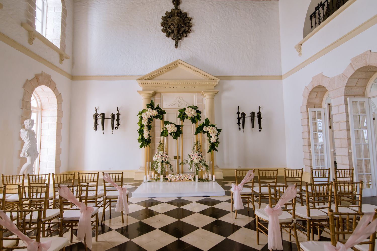 Elegant wedding venue with checkered floors, golden chairs, and ornate architectural details decorated with floral arrangements.