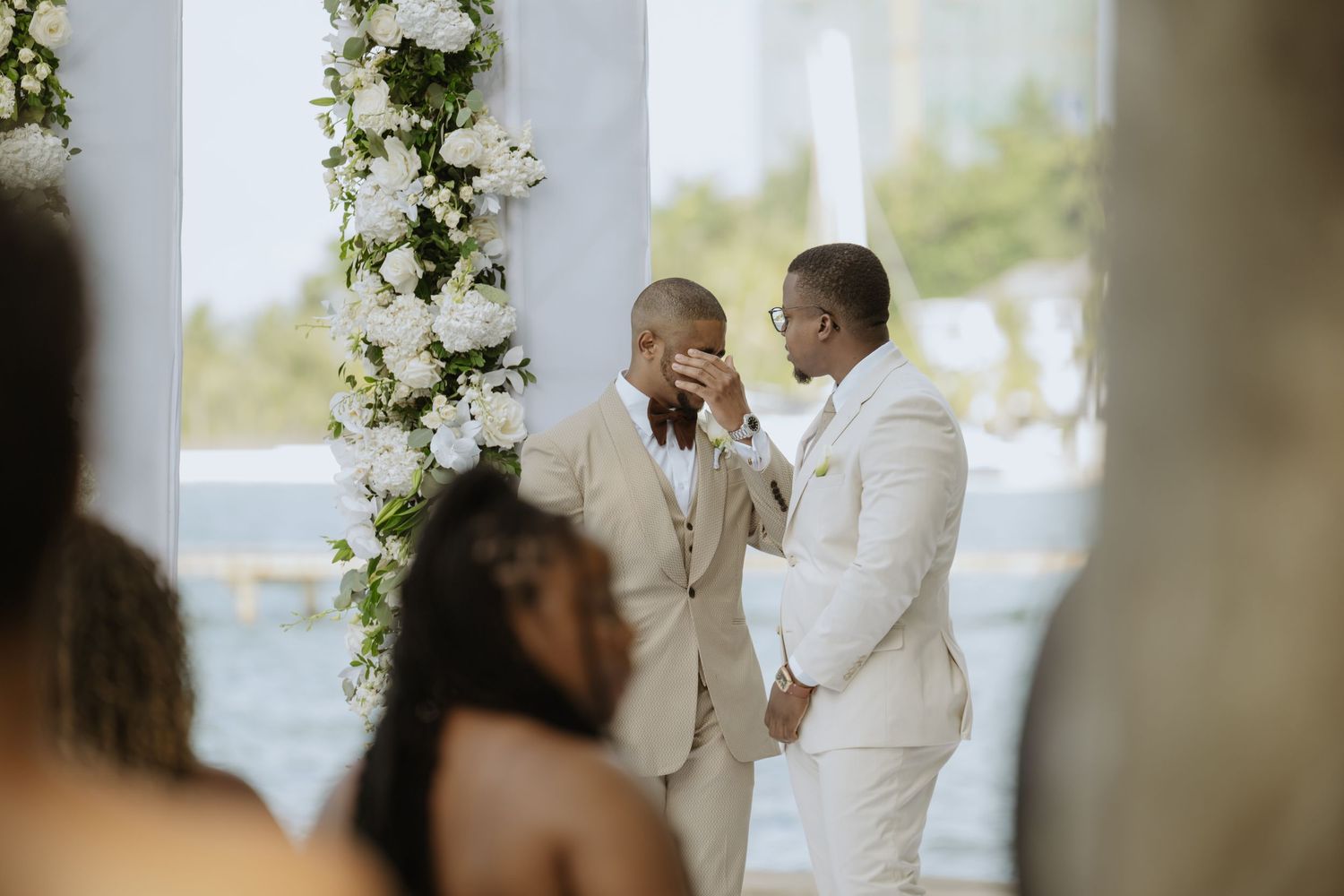 Indoor wedding ceremony with white floral arch and elegant decor by waterfront windows.