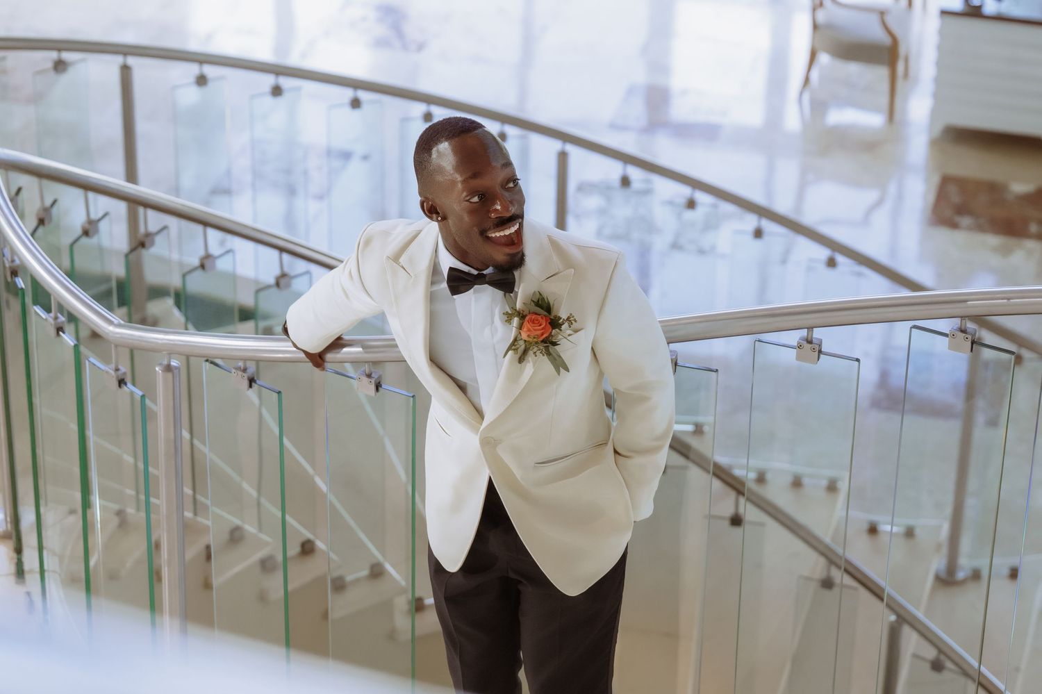 Wedding party member in white suit jacket walking down modern glass-lined staircase.