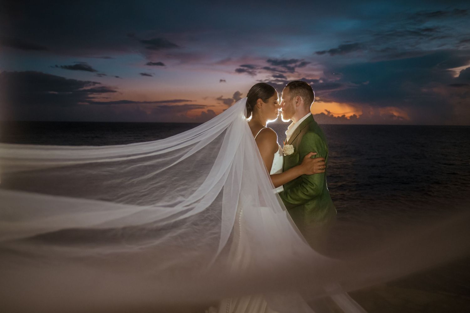 A flowing white wedding veil catches the wind during a dramatic sunset photoshoot on the beach.