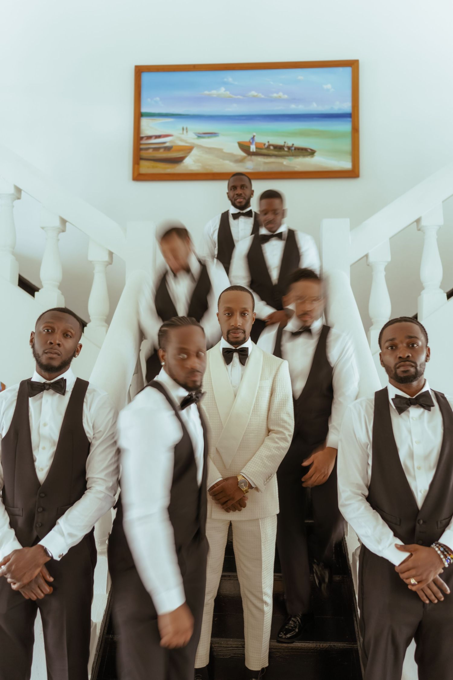 Wedding party members in white suits and black vests pose together against a white wall with framed artwork.