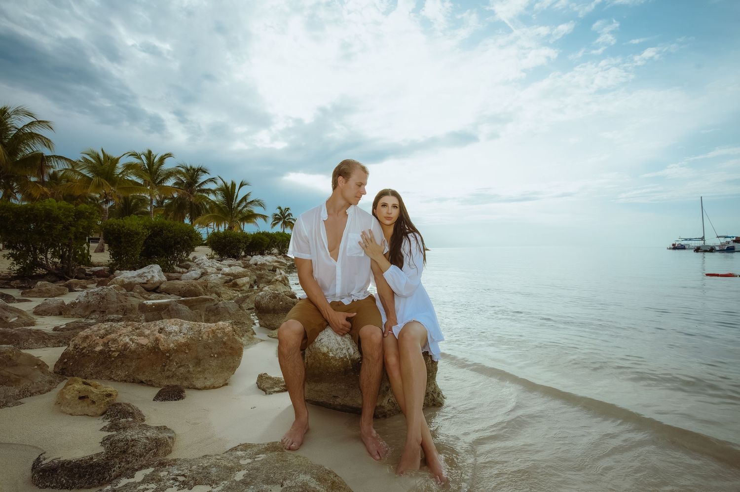 A couple poses together on rocky beach formations with tropical palm trees and boats in the distance.