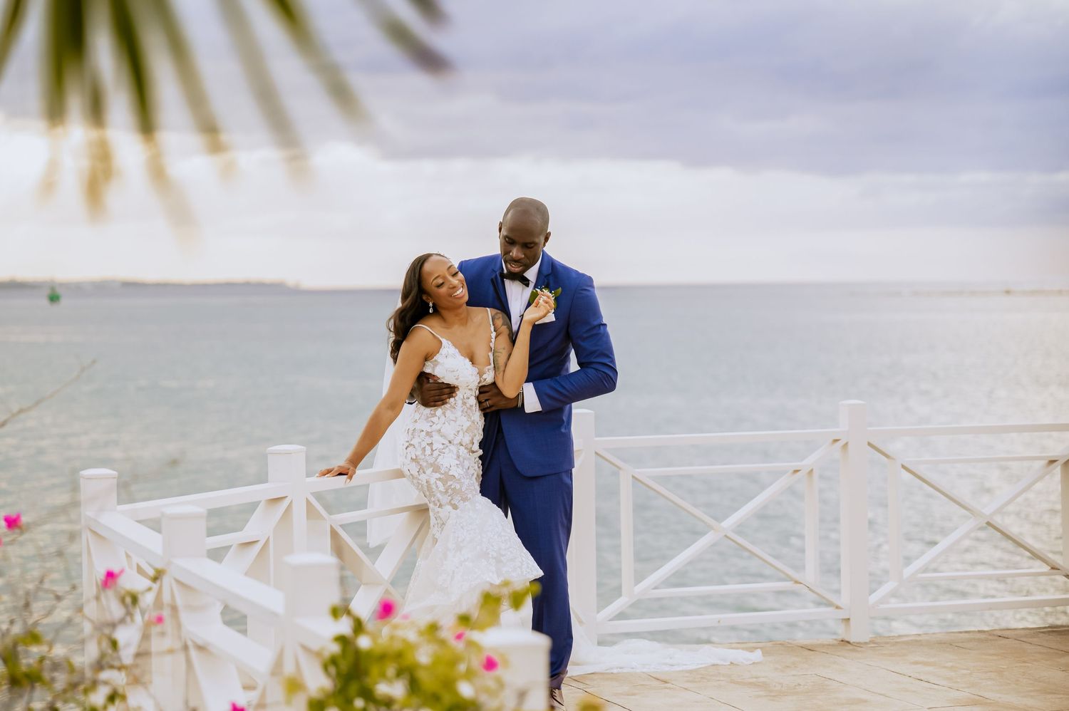A newlywed couple embraces on a tropical beachfront walkway with palm trees in the background.