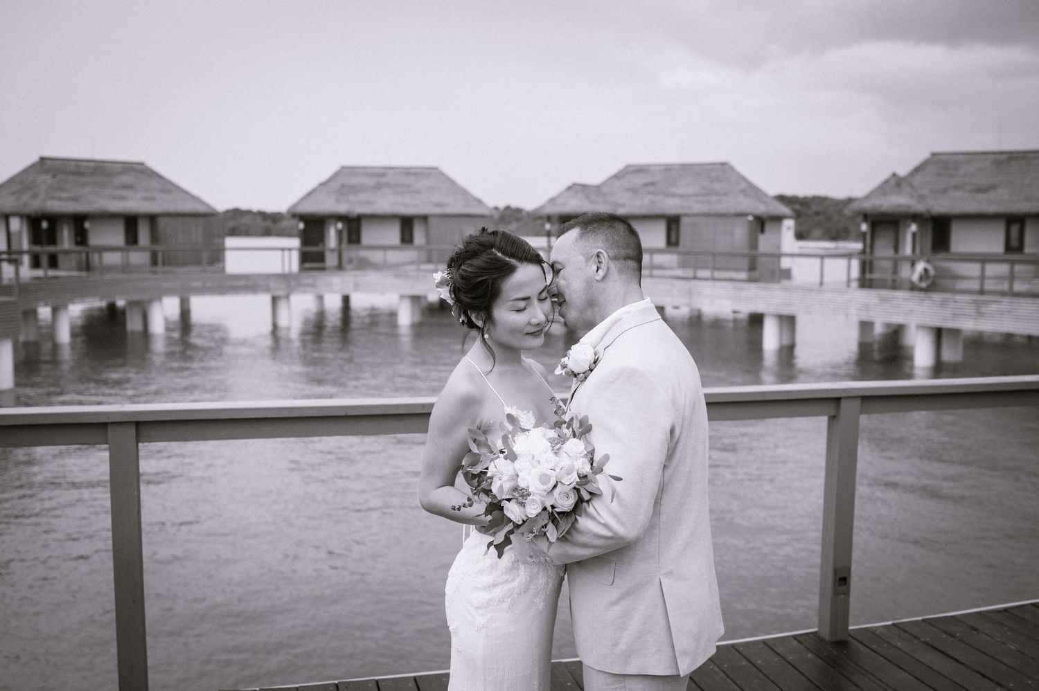 Black and white photo of couple embracing on deck with overwater bungalows behind them.