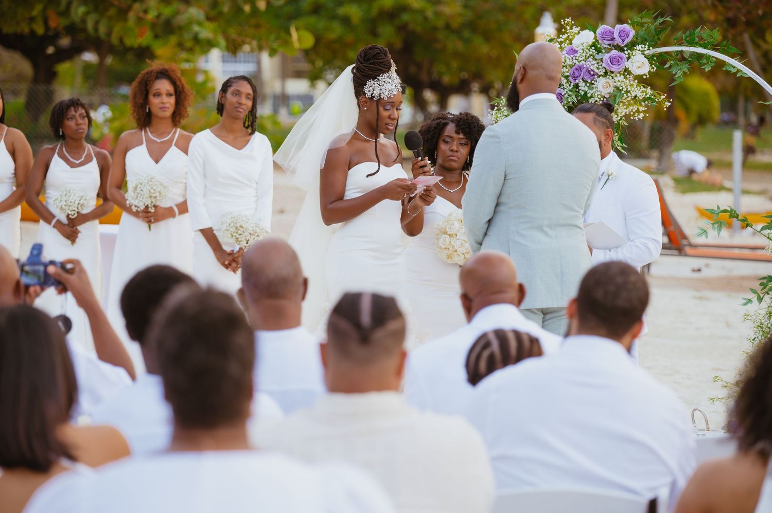 Wedding guests in white gather under tropical trees during an outdoor ceremony.